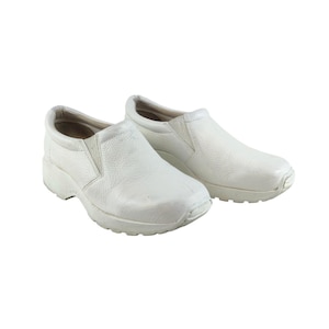 May include: A pair of white slip-on shoes with a textured leather upper. The shoes have a rounded toe and a low, chunky sole. The design features an elastic gore on the top of the foot for easy wear.