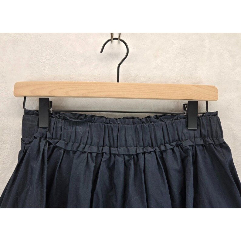 Puede incluir: Una falda azul marino con cintura fruncida y dobladillo con volantes, colgada de una percha de madera con pinzas negras. La falda est&aacute; hecha de una tela suave y fluida.