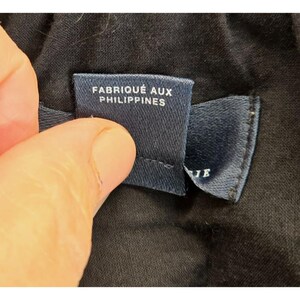 Puede incluir: Primer plano de una etiqueta de tela azul marino con texto blanco que dice "FABRIQU&Eacute; AUX PHILIPPINES". La etiqueta est&aacute; sujeta por una mano y se encuentra sobre un fondo de tela negra.