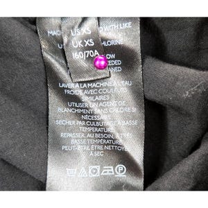 Puede incluir: Primer plano de una etiqueta de tela negra con texto blanco y un bot&oacute;n rosa. La etiqueta incluye instrucciones de lavado en franc&eacute;s e ingl&eacute;s, junto con informaci&oacute;n de tallas: US XS, UK XS y 160/70A.