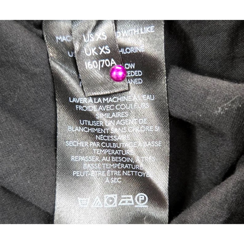 Puede incluir: Primer plano de una etiqueta de tela negra con texto blanco y un bot&oacute;n rosa. La etiqueta incluye instrucciones de lavado en franc&eacute;s e ingl&eacute;s, junto con informaci&oacute;n de tallas: US XS, UK XS y 160/70A.