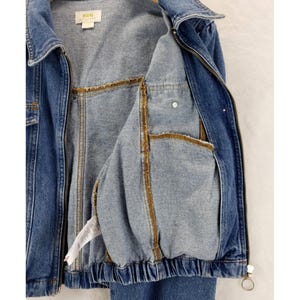 May include: A blue denim jacket with a zip fastening and a collared neckline. The jacket has two front pockets and a light blue interior with gold-coloured stitching. The jacket has a label that reads "MAEVE".
