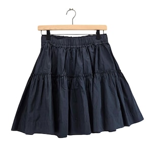 Puede incluir: Falda azul marino con volantes colgada de una percha de madera. La falda tiene una cintura el&aacute;stica y una silueta acampanada. La tela parece ser de algod&oacute;n ligero o similar, con un ligero brillo. La falda est&aacute; dise&ntilde;ada para uso casual.