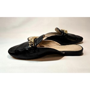 May include: A pair of black leather slip-on shoes with gold-colored chain detailing across the top. The shoes have a rounded toe and a low heel. The background is plain white.