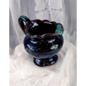 May include: A ceramic pitcher with a dark blue glaze and a scalloped rim. The handle and rim have a teal accent. The pitcher sits on a small pedestal base. The interior is dark, and the exterior has a glossy finish.