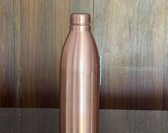 May include: A cylindrical copper water bottle with a smooth, metallic finish and a matching screw-top cap. The bottle features a rounded shoulder design that tapers toward the neck, standing upright with a flat base and a polished, reflective surface.