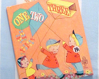 May include: Vintage children's book with an orange cover featuring illustrations of three children flying kites. The kites are labeled "ONE," "TWO," and "THREE." The children wear orange coats, blue pants, and caps. The book has a price tag of 19¢.