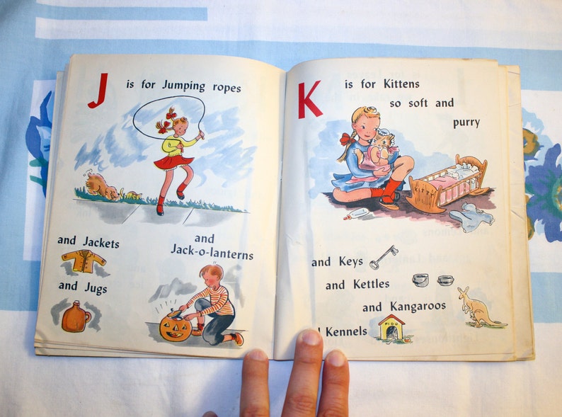 May include: An open children's book with colorful illustrations. The left page features the letter J and the word "Jumping ropes". The right page features the letter K and the word "Kittens".