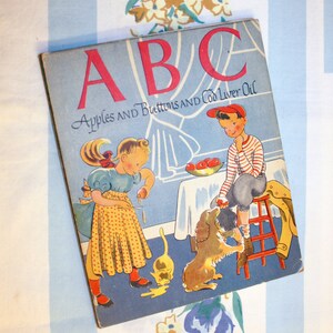 May include: A vintage children's book cover with the title "ABC Apples and Buttons and Cod Liver Oil". The cover features a colorful illustration of two children and a dog in a kitchen setting.