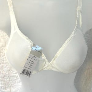 May include: An ivory-colored bra with thin adjustable shoulder straps and a delicate lace trim along the neckline. A product tag attached to the front displays the brand name LA PRIMA, size 80A, and various product codes with a barcode.