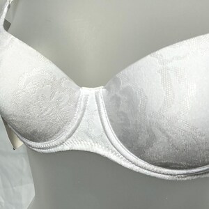 May include: A white underwire bra featuring a tonal floral jacquard pattern across the cups. The garment has thin shoulder straps and a structured design with visible stitching along the bottom edge of the cups and a small tag on the side.