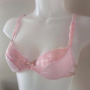 May include: A light pink bra featuring delicate lace detailing on the upper cups and adjustable shoulder straps. The center gore is adorned with a small satin bow and a tiny faux pearl accent, providing a subtle decorative touch to the garment.