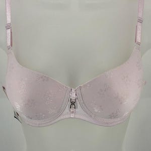 May include: A light pink bra featuring a subtle tonal floral pattern across the cups. The garment includes adjustable shoulder straps and a small silver-toned metal charm detail centered at the bridge between the two cups.