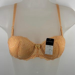 May include: A peach-colored lace bra with thin adjustable straps and a small decorative bow at the center gore. The garment features a structured cup design with intricate floral patterns and includes a black product tag labeled LA VORA.