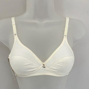 May include: A solid off-white bra featuring adjustable shoulder straps, a soft cup design with horizontal seam detailing across the lower portion of the cups, and a small silver-toned circular charm centered at the bridge of the neckline.