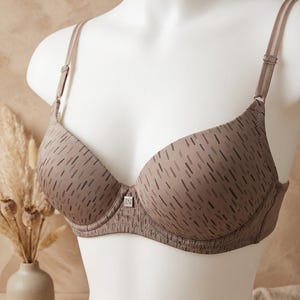 May include: A taupe-colored underwire bra featuring a repeating pattern of dark brown vertical dashes. The bra has adjustable shoulder straps and a small square metal emblem with the letters "LV" centered at the base of the cups.