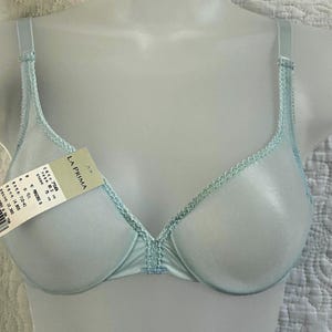 May include: A light blue underwire bra featuring a delicate scalloped lace trim along the neckline and center gore. The garment includes adjustable shoulder straps and a product tag labeled LA PRIMA with size 75B printed on the attached paper tag.