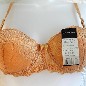 May include: A light orange lace bra with thin adjustable straps and a small center bow. A black and white product tag is attached to the right cup, displaying the brand name LA VORA, size 80A, and various text including 87.5 cm and 80 cm.
