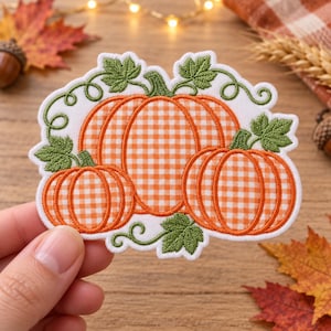 May include: An embroidered patch featuring three pumpkins with an orange and white gingham pattern. The pumpkins are surrounded by green embroidered leaves and curling vines on a white background with a scalloped edge border.