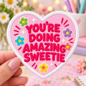 Puede incluir: Un parche bordado en forma de corazón con fondo rosa claro y borde blanco. El texto YOU'RE DOING AMAZING SWEETIE aparece en letras de color rosa oscuro. El diseño incluye pequeñas flores bordadas en verde, azul, morado y amarillo.