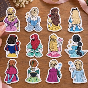 May include: A collection of twelve embroidered patches featuring the backs of various female characters in colourful dresses and hairstyles. Each patch displays intricate thread work depicting distinct outfits, including gowns in shades of purple, blue, yellow, red, green, and pink, with some designs incorporating small star or flower accents.