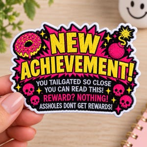 May include: An embroidered patch with a black background featuring a pink donut, a bomb, and four skulls. The text reads: NEW ACHIEVEMENT! YOU TAILGATED SO CLOSE YOU CAN READ THIS! REWARD? NOTHING! ASSHOLES DONT GET REWARDS! It has a white border.