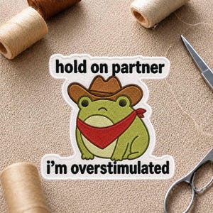 Könnte beinhalten: Ein gestickter Aufnäher mit einem grünen Frosch, der einen braunen Cowboyhut und ein rotes Halstuch trägt. Der Text über dem Frosch lautet hold on partner und der Text darunter lautet i'm overstimulated. Der Aufnäher hat einen weißen Rand.
