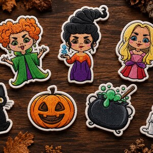 May include: A collection of seven embroidered patches featuring three cartoon characters with distinct hair colours, a black cat, a jack-o'-lantern, a bubbling cauldron with a wooden spoon, and a lit candle, all with white borders on a dark wood surface.