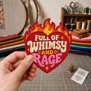 May include: A heart-shaped embroidered patch featuring a flame border in red, orange, and yellow. The text reads FULL OF WHIMSY AND RAGE in cream and pink lettering, accented by small yellow stars. The patch has a red background and stitched edges.