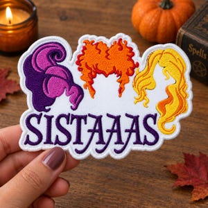 May include: An embroidered patch featuring three stylized hair silhouettes in purple, orange, and yellow, positioned above the purple text "SISTAAAS" on a white background. The patch has a scalloped white border and detailed thread work.