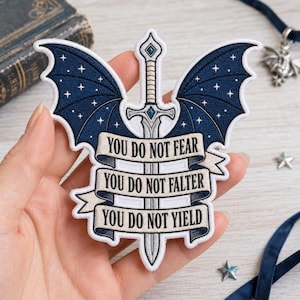 Puede incluir: Un parche bordado que presenta una espada plateada central con detalles en azul, flanqueada por alas de murciélago azul oscuro decoradas con estrellas blancas. Una cinta de color crema sobre la espada muestra el texto: YOU DO NOT FEAR, YOU DO NOT FALTER, YOU DO NOT YIELD.