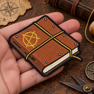 May include: A rectangular embroidered patch featuring a brown book with a yellow anarchy symbol on the cover. The book is secured with yellow straps and has small grey circular accents along the edges, all outlined in a thick black border.