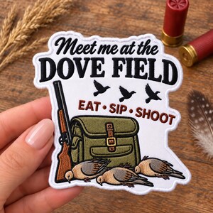 Op de afbeelding: Een witte geborduurde patch met de zwarte tekst Meet me at the DOVE FIELD en bruine tekst EAT SIP SHOOT. Het ontwerp bevat een bruin jachtgeweer, een olijfgroene tas, drie zwarte vogelsilhouetten en drie bruine duiven onderaan.