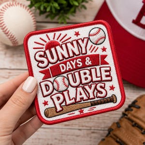 Puede incluir: Un parche bordado cuadrado con fondo blanco y borde rojo. Presenta el texto SUNNY DAYS & DOUBLE PLAYS en rojo y blanco, acompañado de iconos bordados de una pelota de béisbol, un bate de béisbol y un sol, todo en rojo y blanco.