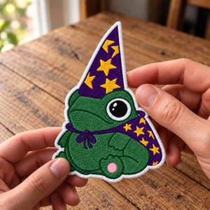 Puede incluir: Un parche bordado que muestra una rana verde con un sombrero de mago y una capa a juego de color morado, ambos decorados con estrellas amarillas. La rana tiene grandes ojos negros con reflejos blancos y una pequeña almohadilla rosa en el pie.