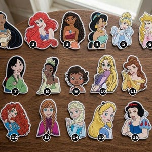 May include: A collection of sixteen embroidered patches featuring various animated female characters, each numbered from 1 to 16. The patches display colorful designs of characters with distinct hairstyles and outfits, arranged in three rows on a wooden surface.