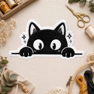 Puede incluir: Un parche bordado que muestra la cabeza de un gato negro con grandes ojos blancos y dos patas negras apoyadas sobre una línea horizontal. El diseño incluye contornos bordados en blanco y pequeños detalles de estrellas de cuatro puntas.