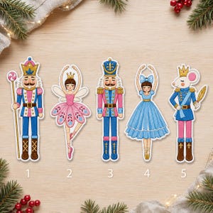 Op de afbeelding: Vijf geborduurde patches met personages in het thema van de Notenkraker: een soldaat met een staf, een fee in een roze tutu, een soldaat in een blauw en roze uniform, een danseres in een blauwe jurk en een muis in een blauw uniform, genummerd één tot vijf.
