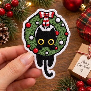 May include: An embroidered patch featuring a black cat with large yellow eyes peeking through a green Christmas wreath decorated with red and white ornaments and a red and white striped bow at the top, finished with a white stitched border.