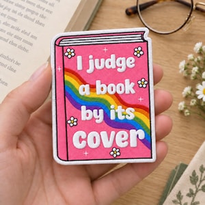 Può includere: Una toppa ricamata rettangolare con il design di una copertina di libro rosa e una striscia arcobaleno che attraversa diagonalmente il centro. Il testo ricamato in bianco recita I judge a book by its cover, con margherite e stelle.