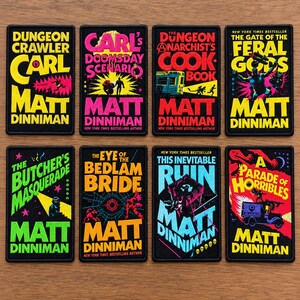 May include: Eight rectangular embroidered patches featuring book cover designs by Matt Dinniman. Titles include Dungeon Crawler Carl, Carl's Doomsday Scenario, The Dungeon Anarchist's Cook Book, The Gate of the Feral Gods, The Butcher's Masquerade, The Eye of the Bedlam Bride, This Inevitable Ruin, and A Parade of Horribles.