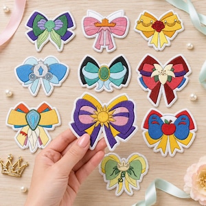 May include: A collection of ten embroidered bow patches featuring various colourful designs, including motifs of a carriage, a red rose, a sun, and an apple. Each patch has a white border and intricate stitching, displayed on a light wood surface.