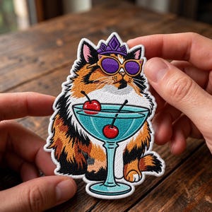 Könnte beinhalten: Ein gestickter Aufnäher mit einer dreifarbigen Katze, die eine violette Krone und eine Sonnenbrille mit violetten Gläsern trägt. Die Katze sitzt hinter einem hellblauen Martiniglas mit zwei roten Kirschen, umrandet von weißem Garn.