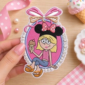 May include: An embroidered patch featuring a cartoon character with blonde hair and black mouse ears, wearing a pink top and blue pants, holding a chocolate ice cream cone. The character is framed by a pink oval with a white border and a pink bow.