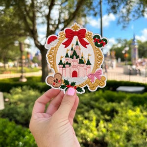 Puede incluir: Un parche bordado que muestra un castillo rosa y verde dentro de un marco ovalado dorado rematado con un lazo rojo. El diseño incluye un gorro de Papá Noel, una corona verde, un icono con forma de ratón marrón, hojas de acebo y un lazo rosa.