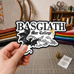 Peut inclure: Un écusson brodé présentant le texte noir BASGIATH et Mar College dans une police gothique. Le motif comprend une silhouette de dragon noir, des nuages blancs stylisés et un petit emblème de dragon doré sur fond blanc avec une bordure noire.