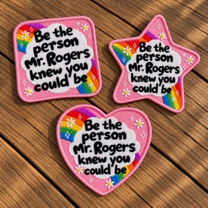 Op de afbeelding: Drie geborduurde patches in vierkante, ster- en hartvorm. Elk heeft een witte wolk in het midden met de zwarte tekst 'Be the person Mr. Rogers knew you could be', omringd door een regenboog, kleine witte madeliefjes en steraccenten op roze.