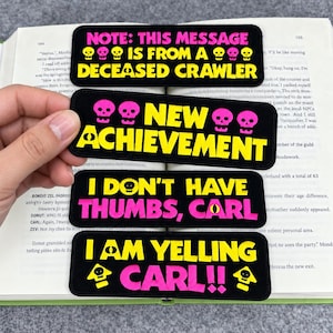 Könnte beinhalten: Vier rechteckige, schwarze, bestickte Aufnäher mit gelbem und pinkfarbenem Text sowie Totenkopf-Symbolen. Die Aufnäher zeigen: NOTE: THIS MESSAGE IS FROM A DECEASED CRAWLER, NEW ACHIEVEMENT, I DON'T HAVE THUMBS, CARL und I AM YELLING CARL!! mit kleinen Symbolen.