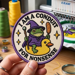 May include: A circular embroidered patch with a cream background and purple border. It features a green frog wearing a purple wizard hat and robe, riding a yellow rubber duck while holding a staff and a corn dog. Text reads: I AM A CONDUIT FOR NONSENSE.