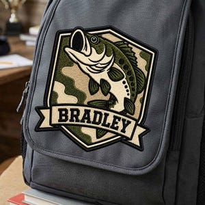 May include: A dark grey backpack featuring a large embroidered patch on the front pocket. The patch displays a green and cream-coloured fish jumping within a shield shape, set against a camouflage background, with the name BRADLEY in black text.